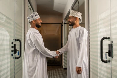 صفقات عمل ناجحة، الخطط الاستراتيجية لتحقيق الأهداف، النقاش و التفاهم بين الموظفين، عقد اجتماع داخل مقر الشركة، مهن و وظائف مكتبية، الاهتمام بالاناقة والمظهر الخارجي، رجلان عربيان خليجيان عمانيان يرتديان الدشداشة و الكمة يصافحان بعضهما البعض في اتفاق
