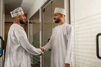 Successful business agreements and deals, strategic plans to achieve goals, discussion and understanding among employees, holding a meeting inside the company headquarters, office professions and jobs, attention to elegance and external appearance, two Arab Gulf men from Oman wearing the dishdasha and the kumma shaking hands with each other.