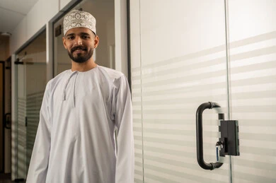 ابواب الكترونية حديثة, ممر طويل لشركة مكتبية بديكور راقي، تصميم عصري حديث لمكاتب الشركات في سلطنة عمان, رجل عربي خليجي عماني مبتسم يرتدي الدشداشة و الكمة يقف بجانب باب زجاجي حديث, مهن و أعمال إدارية
