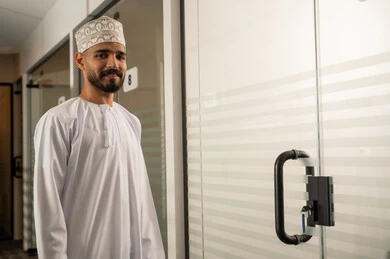 ممر طويل لشركة مكتبية بديكور راقي، تصميم عصري حديث لمكاتب الشركات في سلطنة عمان,  رجل عربي خليجي عماني مبتسم يرتدي الدشداشة و الكمة يقف بجانب باب زجاجي حديث, مهن و أعمال مكتبية إدارية, ابواب الكترونية حديثة من الزجاج