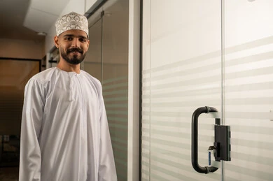 تصميم عصري حديث لمكاتب الشركات في سلطنة عمان, مهن و أعمال مكتبية إدارية, رجل عربي خليجي عماني مبتسم يرتدي الدشداشة و الكمة يقف بجانب باب زجاجي حديث في الشركة, ابواب الكترونية حديثة من الزجاج تصميم عصري حديث لمكاتب الشركات في سلطنة عمان, مهن و أعمال مكتبية إدارية, رجل عربي خليجي عماني مبتسم يرتدي الدشداشة و الكمة يقف بجانب باب زجاجي حديث في الشركة, ابواب الكترونية حديثة من الزجاج