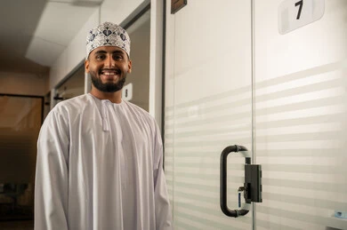 تصميم و ديكور داخلي مكتبي، مهن و أعمال مكتبية إدارية, رجل عربي خليجي عماني مبتسم يرتدي الدشداشة و الكمة يقف بجانب باب زجاجي حديث في الشركة, ابواب الكترونية حديثة من الزجاج