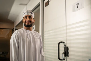Modern electronic glass doors, a long corridor for an office company with elegant decor, a modern contemporary design for corporate offices in the Sultanate of Oman, a smiling Arab Gulf Omani man wearing a dishdasha and kumma standing next to a modern glass door, administrative office professions and jobs.