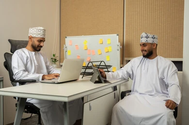 Dialogue and discussion and providing services to customers, friendliness and good reception, a comfortable work environment in upscale companies, the concept of cooperation and teamwork, expressions and gestures of happiness and joy, two smiling Arab Gulf Omani men wearing the dishdasha and kumma in the office and using electronic devices.