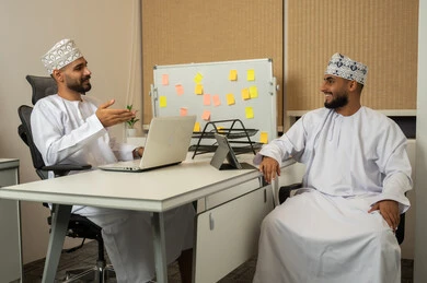 بيئة العمل المريحة في الشركات الراقية، الحوار والنقاش وتقديم الخدمات للعملاء، البشاشة وحسن الاستقبال, مفهوم التعاون والعمل المشترك, تعابير وايماءات السعادة والسرور، رجلان عربيان خليجيان عمانيان مبتسمان يرتديان الدشداشة و الكمة في المكتب و يقومان باستخدام الأجهزة الالكترونية