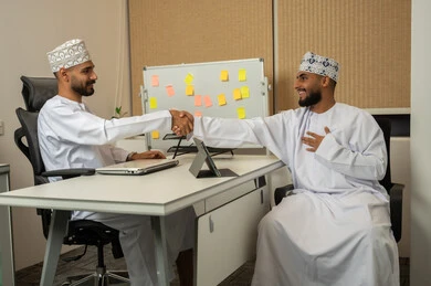 مفهوم الترحيب و السلام و إلقاء التحية، بيئة العمل المريحة في الشركات الراقية، مفهوم التعاون والعمل المشترك, تعابير وايماءات السعادة والسرور، رجلان عربيان خليجيان عمانيان مبتسمان يرتديان الدشداشة و الكمة يجلسان في المكتب و يقومان بمصافحة بعضهم, استخدام الأجهزة المتطورة