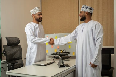 A handshake and warm greeting between two men, a comfortable work environment in upscale companies, the concept of collaboration and teamwork, expressions and gestures of happiness and joy, two smiling Arab Gulf men from Oman wearing the dishdasha and kumma standing in front of the office and shaking hands with each other, using advanced devices.