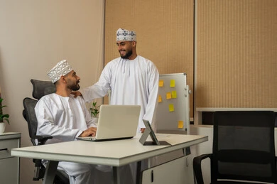 Moral motivation among colleagues, exchanging experiences and information to achieve goals, developing and studying new projects in Omani companies, gestures of happiness and joy, the concept of cooperation and joint work, two smiling Arab Gulf men from Oman wearing the dishdasha and kumma, with one patting the shoulder of the other.