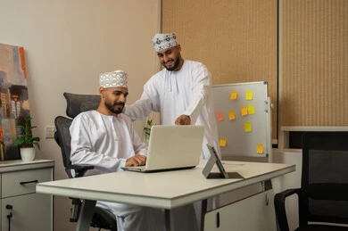 Development and study of new projects in Omani companies, discussing ideas and exchanging opinions, gestures of happiness and joy, the concept of cooperation and joint work, two smiling Arab Gulf men from Oman wearing the dishdasha and kumma, one sitting at the desk in front of the laptop and the other pointing to the computer.