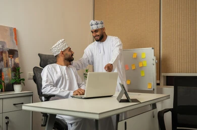 Exchange of experiences and information to achieve goals, developing and studying new projects in Omani companies, gestures of happiness and joy, the concept of cooperation and joint work, two smiling Arab Gulf Omani men wearing the dishdasha and kumma, one sitting at the desk in front of the laptop and the other pointing to the computer.