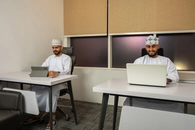 Monitoring and developing work, understanding and cooperation among colleagues to achieve the company's goals, the concept of business management, completing tasks using modern devices. Two smiling Arab Gulf men from Oman, wearing the dishdasha and kumma, are sitting in the office and working on a laptop and a tablet.