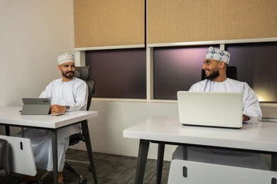 Understanding and cooperation among colleagues to achieve the company's goals, the concept of business management, completing tasks through modern devices, following up on the work strategy, two smiling Arab Gulf men from Oman wearing the dishdasha and kumma sitting in the office and working on a laptop and tablet.