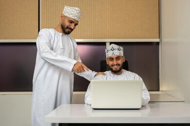 الحوار و تبادل الأفكار، ايماءات السعادة و السرور، الاشارة الي شاشة الحاسوب المحمول, مفهوم التعاون والعمل المشترك, رجلان عربيان خليجيان عمانيان مبتسمان يرتديان الدشداشة و الكمة يجلس أحدهما على المكتب أمام اللابتوب و يشير الآخر الى شاشة الكمبيوتر