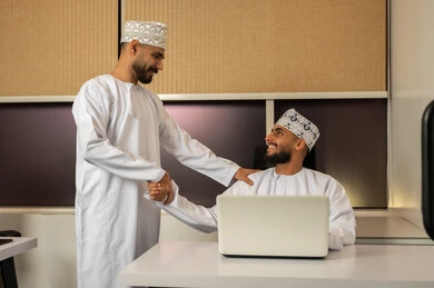الترحيب و السلام و إلقاء التحية، مفهوم التعاون والعمل المشترك, تعابير وايماءات السعادة والسرور، رجلان عربيان خليجيان عمانيان مبتسمان يرتديان الدشداشة و الكمة يجلس أحدهما على المكتب أمام اللابتوب و يصافح الاخر