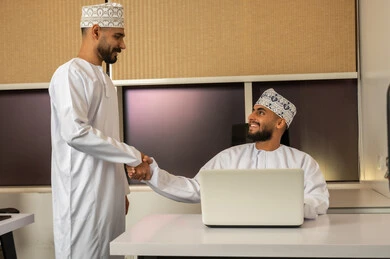 المصافحة و الترحيب و الاستقبال, مفهوم التعاون والعمل المشترك, تعابير وايماءات السعادة والسرور، رجلان عربيان خليجيان عمانيان مبتسمان يرتديان الدشداشة و الكمة يجلس أحدهما على المكتب أمام اللابتوب و يصافح الاخر