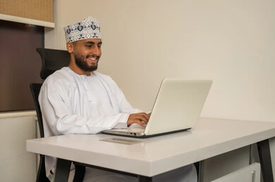 الكتابة عبر النقر على الكيبورد، دمج العمل مع التقنية والتكنولوجيا، إدارة شؤون الأعمال وتنظيمها عبر الأجهزة التقنية، رجل عربي خليجي عماني يرتدي الدشداشة و الكمة يقوم باستخدام اللابتوب, مفهوم إنجاز المهام عن بعد, بيئة العمل المكتبي المريح