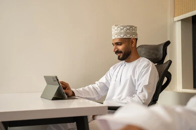 بيئة العمل المكتبي المريح, متابعة مواقع التواصل الاجتماعي،  دمج التقنيات الحديثة و المتطورة مع العمل, مهن و أعمال مكتبية إدارية, رجل عربي خليجي عماني مبتسم يرتدي الدشداشة و الكمة يجلس على المكتب امام جهاز التابلت, تعابير وايماءات الاندماج