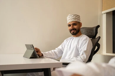 Project management professionally using a tablet, a comfortable office work environment, monitoring social media, using modern and advanced technologies, administrative office jobs and professions, a smiling Arab Gulf Omani man wearing a dishdasha and kumma sitting at a desk in front of a tablet, looking at the camera with gestures of happiness and joy.