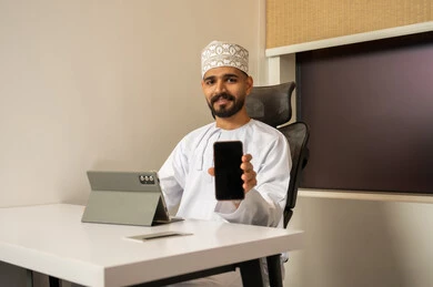 Performing tasks via mobile, enjoying exchanging conversations and news, integrating modern and advanced technologies with work, professions and administrative office work, a smiling Arab Gulf Omani man wearing a dishdasha and kumma sitting at a desk in front of a tablet and displaying the mobile screen.