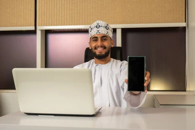 Preparing and reviewing performance reports, business feasibility studies, using a laptop to send emails, integrating modern and advanced technologies with work, administrative office professions and jobs, a smiling Arab Gulf Omani man wearing a dishdasha and kumma sitting at a desk in front of a computer and displaying a mobile screen.