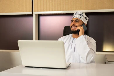 Using a laptop to send emails, communicating with clients remotely, integrating modern and advanced technologies with work, professions and administrative office jobs, a smiling Arab Gulf Omani man wearing a dishdasha and kumma sitting at a desk in front of a computer and talking on a mobile phone, expressions and gestures of integration.