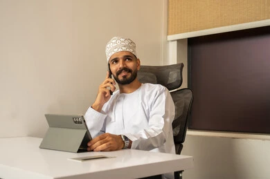 مهن و أعمال مكتبية إدارية, رجل عربي خليجي عماني مبتسم يرتدي الدشداشة و الكمة يجلس على المكتب امام جهاز التابلت و يتحدث عبر المحمول, تعابير وايماءات الاندماج, التواصل مع العملاء عن بعد، دمج التقنيات الحديثة و المتطورة مع العمل
