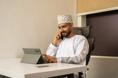 التواصل مع العملاء عن بعد، دمج التقنيات الحديثة و المتطورة مع العمل, مهن و أعمال مكتبية إدارية, رجل عربي خليجي عماني مبتسم يرتدي الدشداشة و الكمة  يجلس على المكتب امام جهاز التابلت, تعابير وايماءات الاندماج
