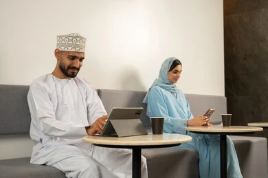 A work session in the café, using modern technologies, enjoying hot beverages, a smiling Omani Gulf Arab man wearing a dishdasha and kumma using a tablet, and next to him, a veiled Omani Gulf Arab woman wearing a blue abaya using her phone to complete tasks remotely.