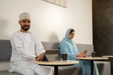 Completing the required tasks, enjoying favorite drinks, using modern technologies, a smiling Omani Gulf Arab man wearing a dishdasha and kumma using a tablet, next to him is a veiled Omani Gulf Arab woman wearing a blue abaya and using a phone, working online at the café.
