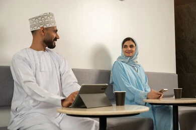 Use modern technologies, enjoy drinking hot beverages, socializing and exchanging conversations, a smiling Omani Gulf Arab man wearing a dishdasha and kumma using a tablet, next to him is a veiled Omani Gulf Arab woman wearing a blue abaya and using a phone, working remotely in a café.