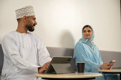 انجاز مهام العمل عن بعد، استخدم التقنيات الحديثة,  الاستمتاع بتناول المشروب الساخن, التسامر وتبادل الاحاديث, رجل عربي خليجي عماني مبتسم يرتدي الدشداشة و الكمة يستخدم جهاز التابلت و بجانبه سيدة عربية خليجية عمانية محجبة ترتدي عباءة زرقاء تستخدم الهاتف و تجلس في المقهى