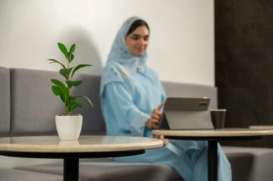 قضاء وقت ممتع في الخارج، ايماءات التركيز والسرور، العمل عن بعد، إنجاز المهام اليوميه، دمج التقنية والتكنولوجيا في الروتين اليومي، الاستمتاع بتناول المشروبات الساخنة, سيدة عربية خليجية عمانية مبتسمة محجبة ترتدي عباءة زرقاء و تجلس في مقهى و تستخدم جهاز التابلت