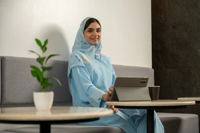 مفهوم العمل عن بعد، دمج التقنية و التكنولوجيا في الروتين اليومي، النظر الى الكاميرا بسعادة, سيدة عربية خليجية عمانية مبتسمة محجبة ترتدي عباءة زرقاء و تجلس في مقهى, امرأة سعودية تستخدم جهاز التابلت و تستمتع بتناول مشروب ساخن