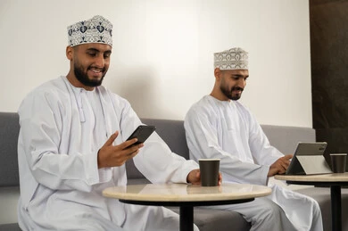 Browsing social media, expressions and gestures of integration and joy, two smiling Arab Gulf men from Oman wearing the dishdasha and kumma sitting in a café, using modern technological devices, enjoying a hot drink, working remotely.