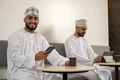 النظر الى الكاميرا بايماءات السعادة, تصفح مواقع التواصل الاجتماعي، رجلان عربيان خليجيان عمانيان مبتسمان يرتديان الدشداشة و الكمة و يجلسان في مقهى, استخدام الأجهزة التكنولوجية الحديثة, الاستمتاع بتناول المشروبات الساخنة, العمل عن بعد