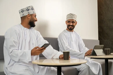 Socializing and exchanging conversations, two smiling Arab Gulf Omani men wearing dishdasha and kumma sitting in a café, using modern technological devices, enjoying hot beverages, working remotely.