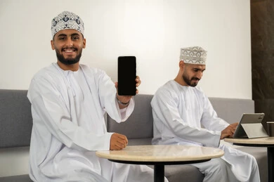 عرض شاشة الجوال,  رجلان عربيان خليجيان عمانيان مبتسمان يرتديان الدشداشة و الكمة و يجلسان في مقهى, استخدام الأجهزة التكنولوجية الحديثة, العمل عن بعد, النظر الى الكاميرا بايماءات السعادة