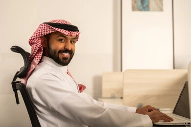 Completion of projects with precision, designated spaces for each employee in the company, focus and meticulous work, business and company development, discussing plans and explaining job tasks, an Arab Gulf Saudi employee wearing traditional attire and a ghutrah using a laptop.