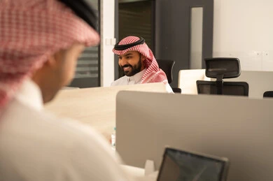 Dedicated spaces for each employee in the company, focus and meticulous work, dialogues and discussions within the team, business and company development, work meetings and project completion, discussing plans and explaining work tasks, Arab Saudi Gulf colleagues wearing traditional attire and the ghutrah using a tablet.