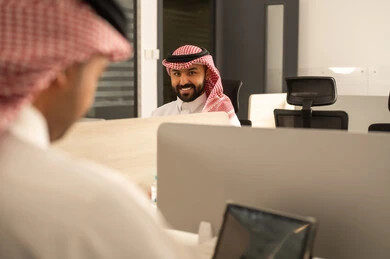 Dedicated spaces for each employee in the company, focus and meticulous work, dialogues and discussions within the team, business and company development, work meetings and project completion, discussing plans and explaining work tasks, Arab Saudi Gulf colleagues wearing traditional attire and the ghutrah using a tablet.