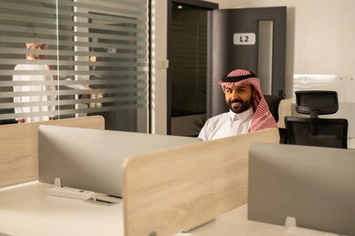 Designated spaces for each employee in the company, focus and meticulous work, business and company development, completing projects accurately, discussing plans and explaining work tasks, an Arab Gulf Saudi employee wearing traditional attire and a ghutrah using a laptop.