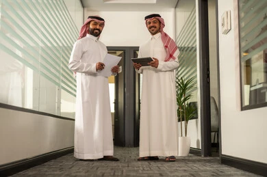 Two Saudi Arabian Gulf Arab employees are sitting inside the office, discussing project plans, using a tablet and a laptop to complete tasks, focusing and working meticulously, engaging in dialogues and discussions within the team, developing businesses and companies, and explaining job responsibilities.