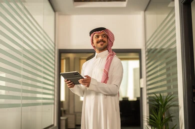 The concept of business management and achieving the company's goals, an Arab Gulf Saudi businessman wearing traditional attire and a ghutrah working on a tablet, office professions and jobs, completing work tasks remotely.