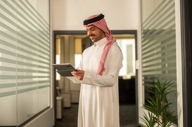 Office professions and jobs, the concept of business management and achieving company goals, a Gulf Arab businessman from Saudi Arabia wearing traditional attire and a ghutrah working on a tablet, accomplishing remote work tasks.