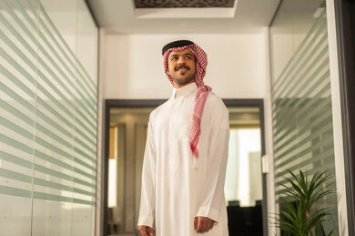 The evolution in the design of offices and upscale companies, working in the administrative and office sector, professions and jobs in Saudi Arabia, an Arab Gulf Saudi man wearing traditional attire and a ghutrah stands next to glass windows overlooking the towers and skyscrapers, looking at the camera happily.
