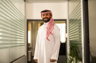رجل عربي خليجي سعودي يرتدي الزى التقليدي والشماغ يقف بجانب النوافذ الزجاجية المطلة على الأبراج وناطحات السحاب وينظر إلى الكاميرا بسعادة، التطور في تصميم المكاتب والشركات الراقية، العمل في القطاع الإداري والمكتبي، المهن والوظائف في السعودية