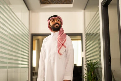 The evolution in the design of offices and upscale companies, working in the administrative and office sector, professions and jobs in Saudi Arabia, an Arab Gulf Saudi man wearing traditional attire and a ghutrah stands next to glass windows overlooking the towers and skyscrapers, looking at the camera happily.