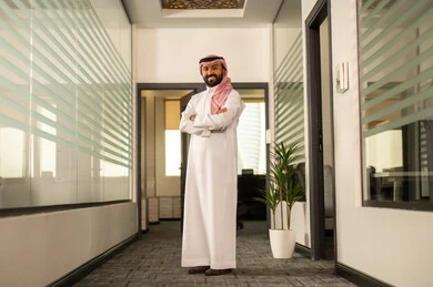 التطور في تصميم المكاتب والشركات الراقية، العمل في القطاع الإداري والمكتبي، المهن والوظائف في السعودية، رجل عربي خليجي سعودي يرتدي الزى التقليدي والشماغ يقف بجانب النوافذ الزجاجية المطلة على الأبراج وناطحات السحاب وينظر إلى الكاميرا بسعادة