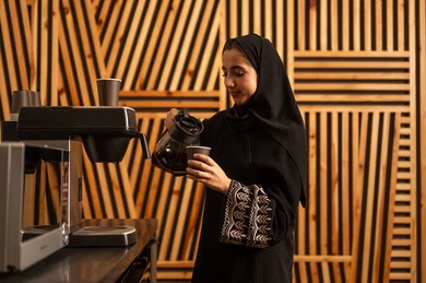 امراه عربيه خليجيه سعوديه ترتدي العباءه والحجاب تستخدم اله تحضير القهوة السريعة، منتج وطني محلي سعودي، الاستمتاع باعداد المشروبات الساخنة الطازجة، مهن ووظائف نسائية ، العمل في القطاع الإداري والمكتبي، غرفة استراحة العمل