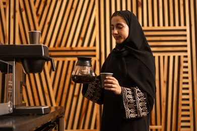 Women's professions and jobs, working in the administrative and office sector, break room, an Arab Gulf Saudi woman wearing an abaya and hijab using a quick coffee maker, a local Saudi national product, enjoying preparing fresh hot beverages. Women's professions and jobs, working in the administrative and office sector, break room, an Arab Gulf Saudi woman wearing an abaya and hijab using a quick coffee maker, a local Saudi national product, enjoying preparing fresh hot beverages.